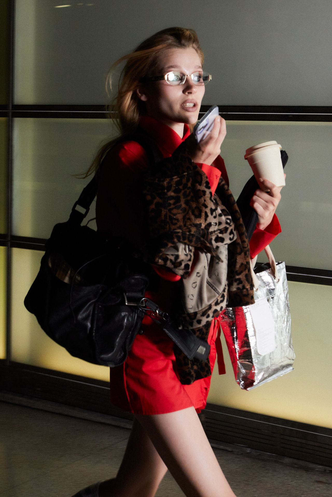 Woman in a red shirt and leopard-print coat carrying bags and a coffee while checking her phone